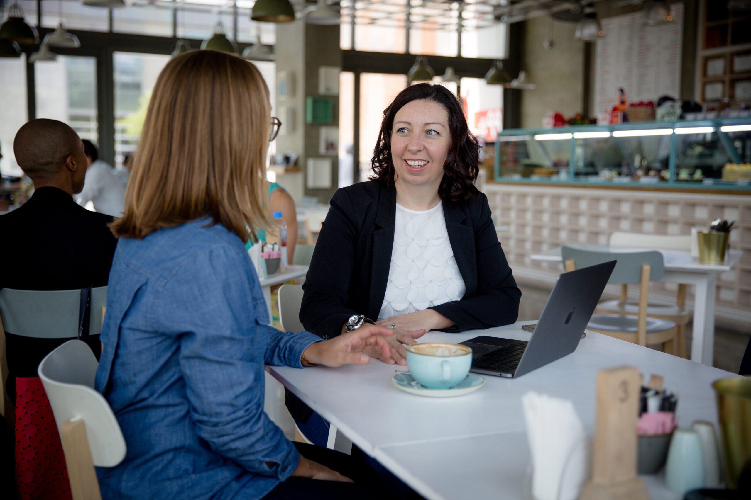 Two women sitting next to each other at a cafe table talking. There are coffee cups and a laptop on the table and the dark haired women looks to be speaking.